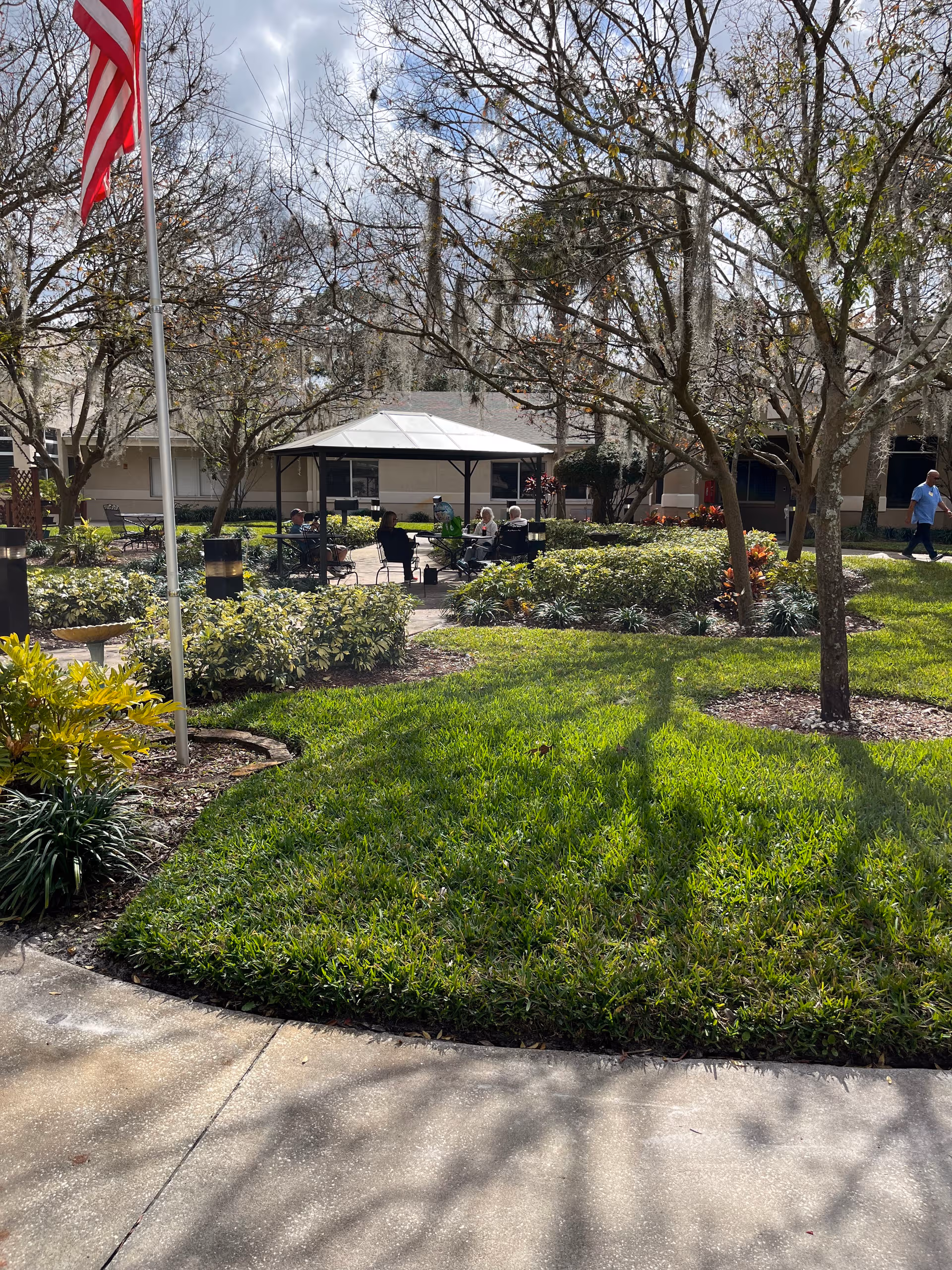 Outdoor courtyard with a shaded seating area; residents sit around tables amid trees and shrubs.