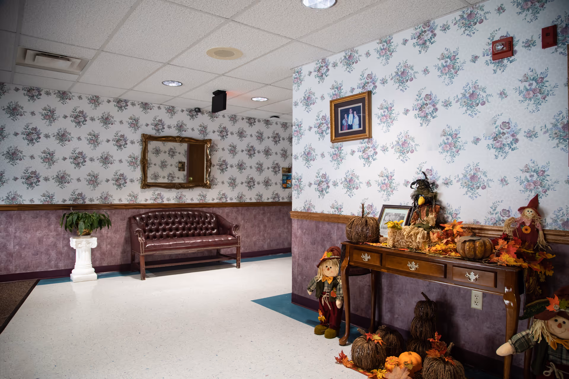 Interior lobby area with floral wallpaper, purple tufted sofa, and autumn decor on a console.