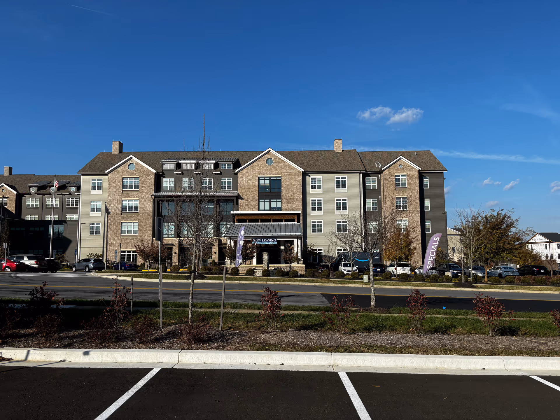 Front facade of a multi-story senior living building with a parking area and landscaped median under a clear blue sky.