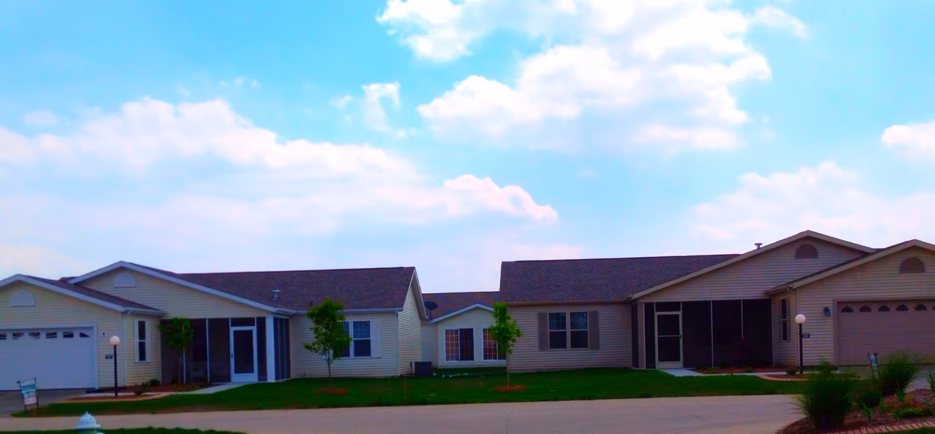 Exterior view of a single-story residential building with beige siding, multiple garage doors, screened porch areas, small trees, and a well-maintained lawn under a partly cloudy sky.