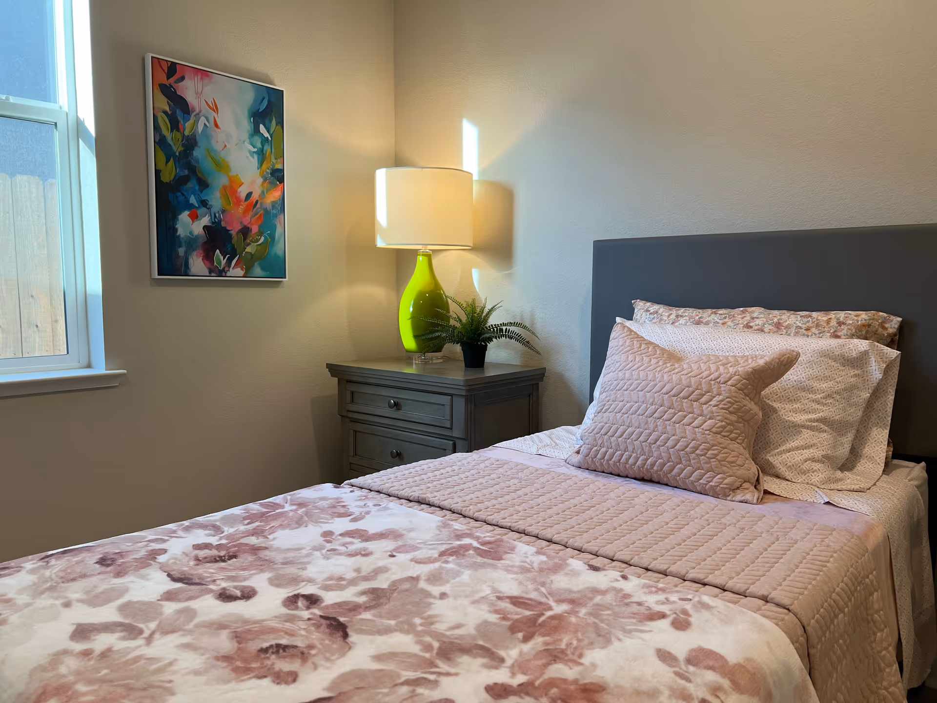A neatly made bedroom featuring a floral quilt, pillows, lamp, nightstand, and wall art.