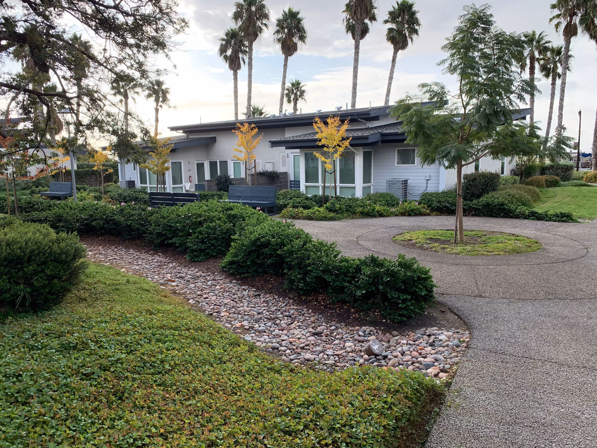 Low-rise residential building front with landscaped paths, shrubs, palm trees and benches under a cloudy sky.