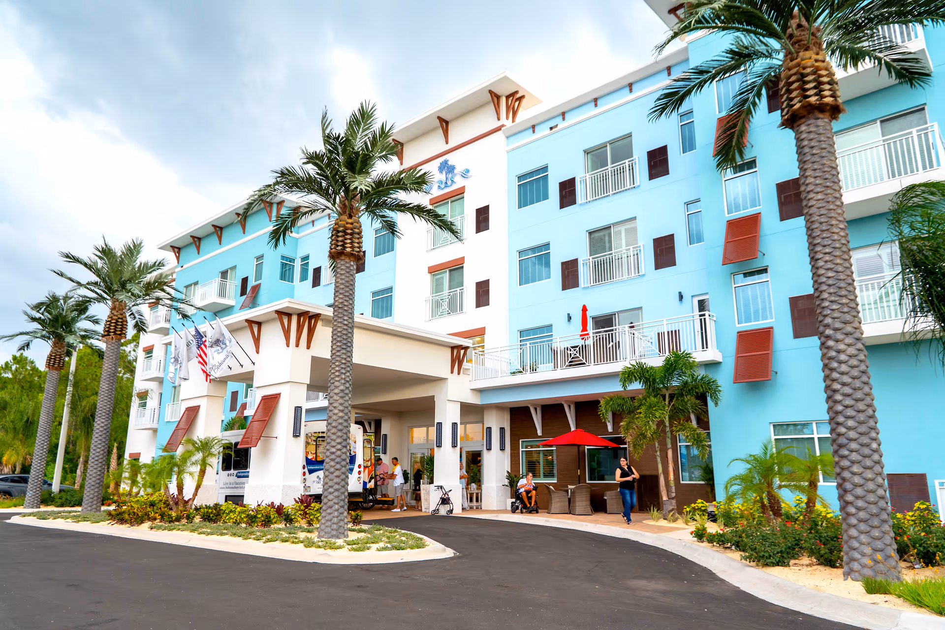 Exterior view of a multi-story senior living facility with a light blue facade, palm trees lining the driveway, and a covered entrance where people are gathered near a shuttle bus. There are balconies with railings and red awnings on some windows.