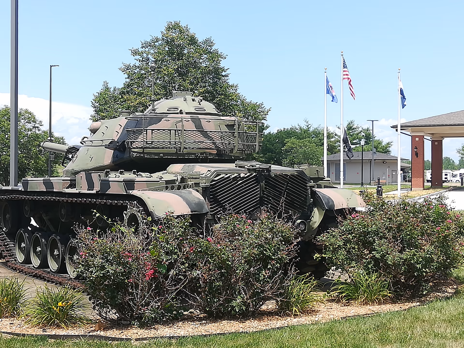 A camouflaged tank displayed outdoors near flags and a building at a veterans home.