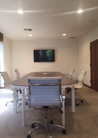 A conference room with a large oval table, white chairs, and a wall-mounted TV.