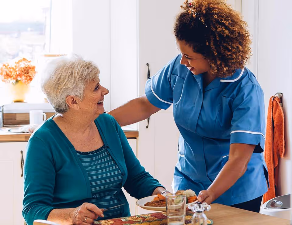 Caregiver in blue uniform assists an elderly woman during a meal at a dining table.