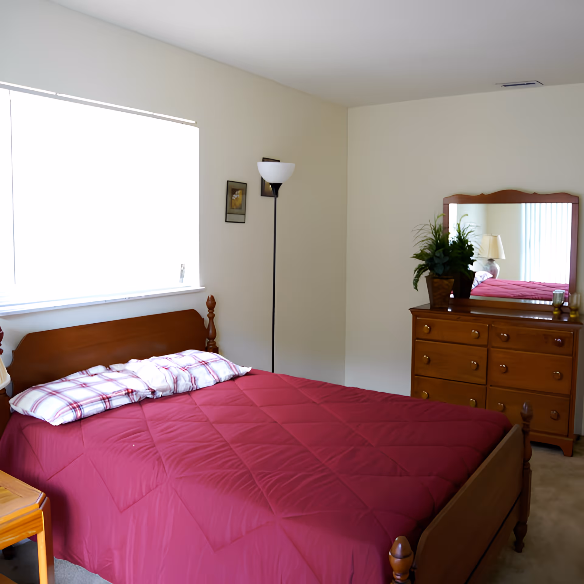 A bedroom with a wooden bed frame and a red quilted bedspread. There are two pillows with white and red plaid pillowcases. A wooden dresser with a mirror on top is against the wall, decorated with a potted plant and a lamp reflected in the mirror. A tall floor lamp stands in the corner next to two small framed pictures on the wall. A window with blinds is on the left side, letting in bright natural light.