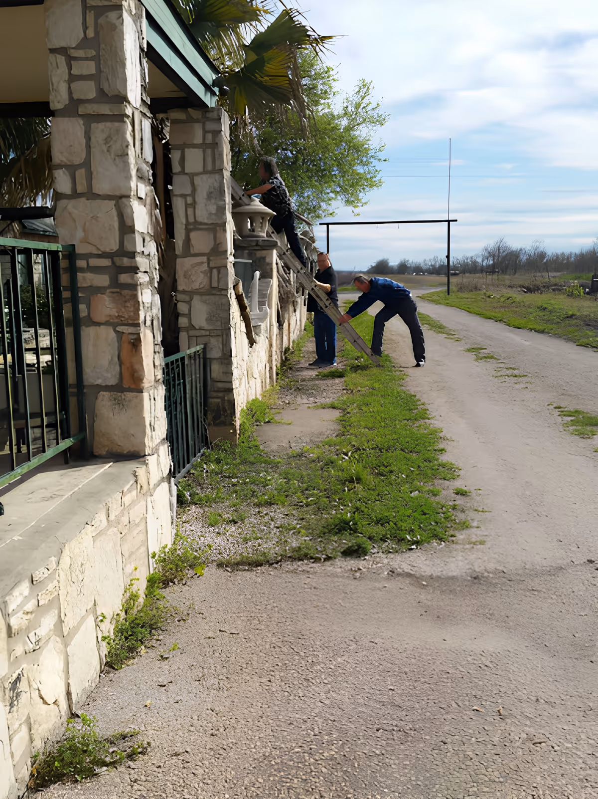 Three people set up a ladder against a stone exterior wall next to a rural dirt road.