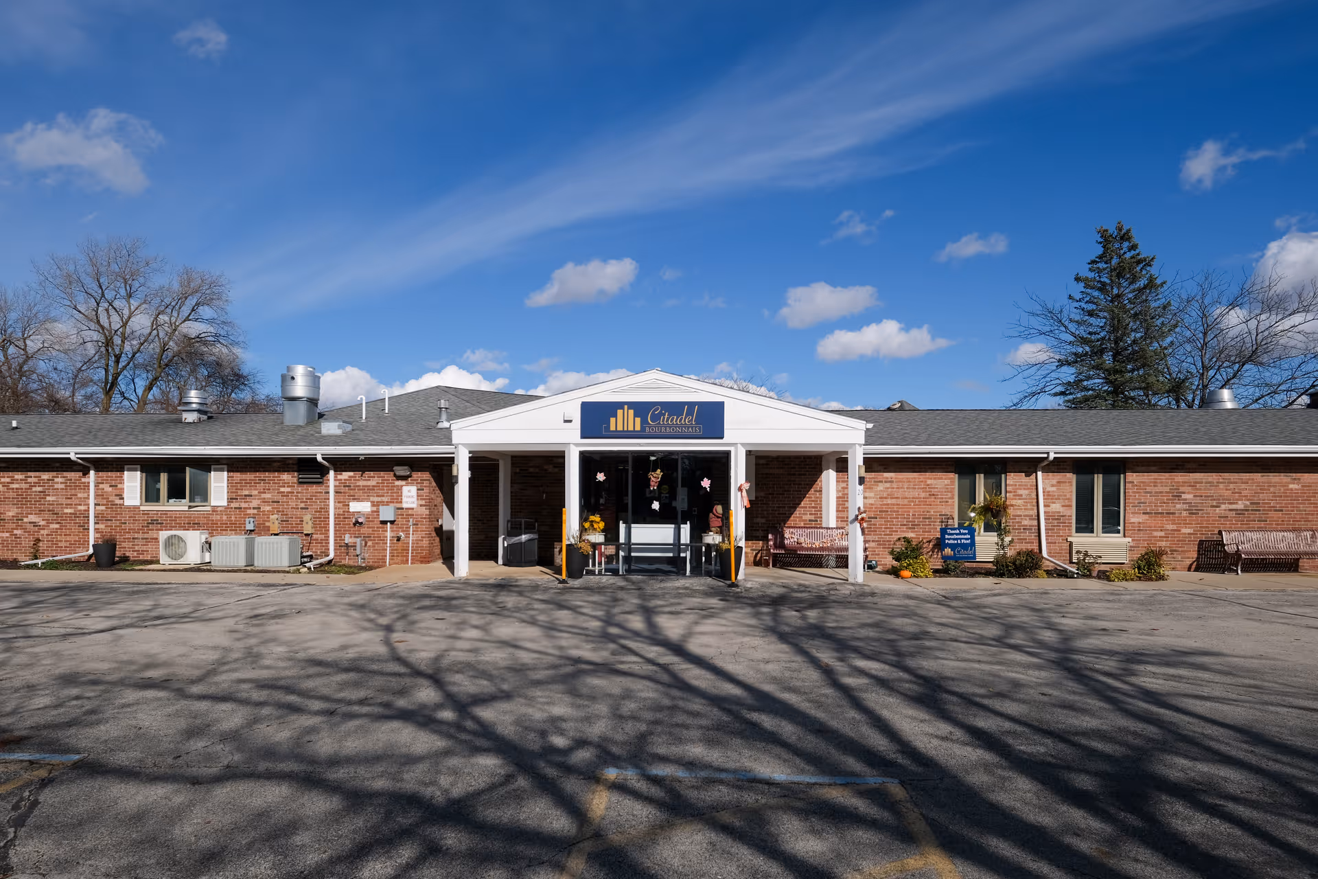 Front exterior view of the Citadel of Bourbonnais facility, a single-story brick building with a covered entrance and a sign above the door. The sky is clear with a few clouds, and there are benches and some plants near the entrance.