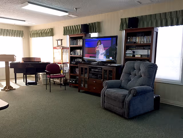 A cozy living room area with a large gray recliner chair, a TV on a wooden stand displaying a woman speaking, two wooden bookshelves filled with DVDs and books, a maroon chair, a piano, and a popcorn machine near windows with green valances.