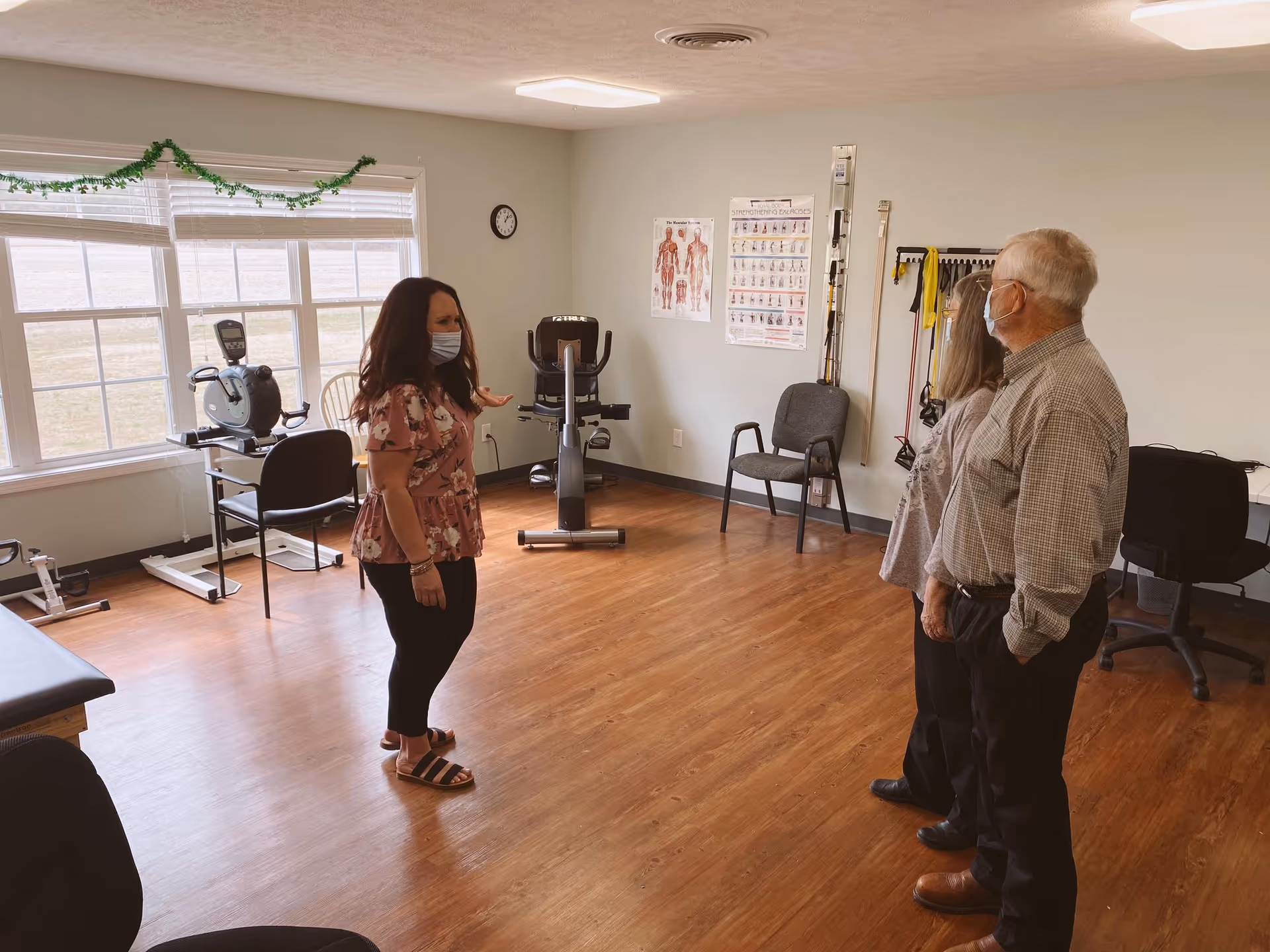 A woman wearing a mask stands in a room with wooden flooring, talking to an elderly man and woman who are also wearing masks. The room has exercise equipment, chairs, and anatomical posters on the wall, suggesting a physical therapy or fitness area.