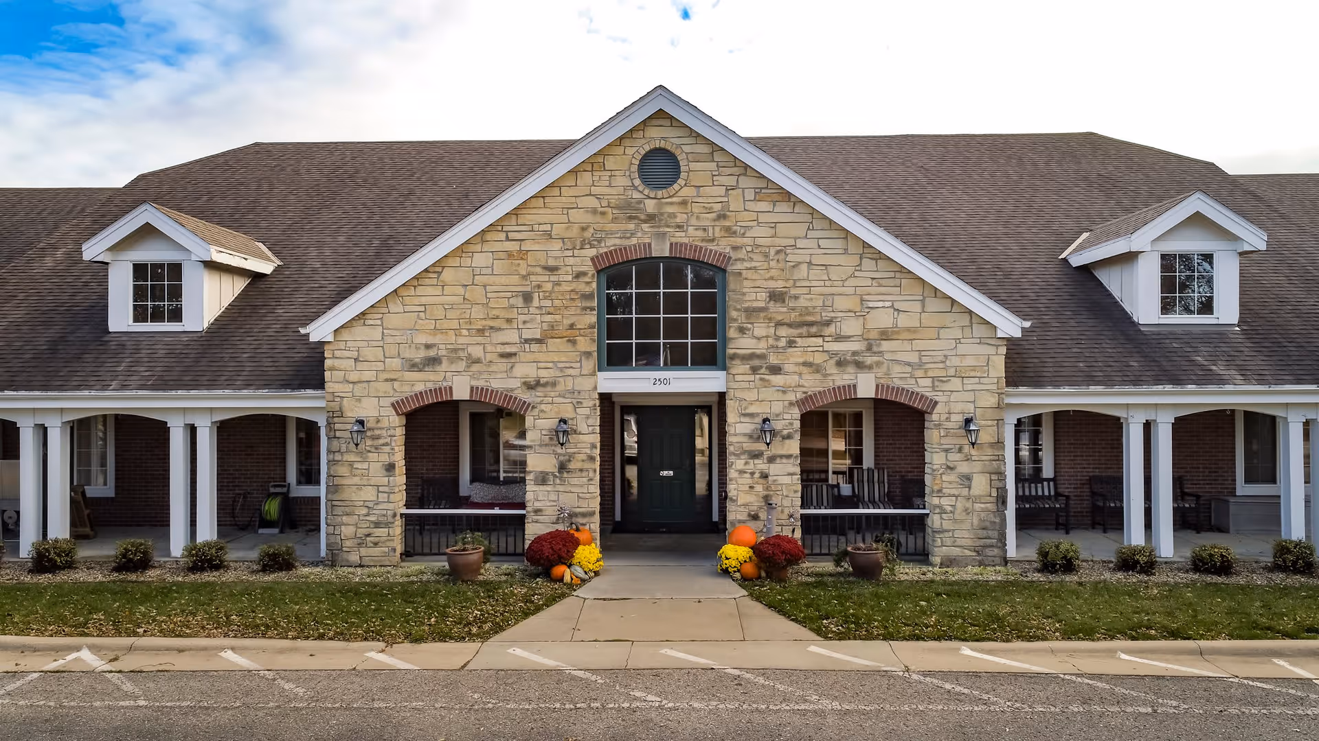 Front exterior view of a single-story assisted living facility building with a stone facade and a dark shingled roof. The entrance is centered with a large window above the door and two archways on either side. There are potted plants and fall decorations near the entrance. The building has white columns supporting a covered porch area on both sides of the entrance.
