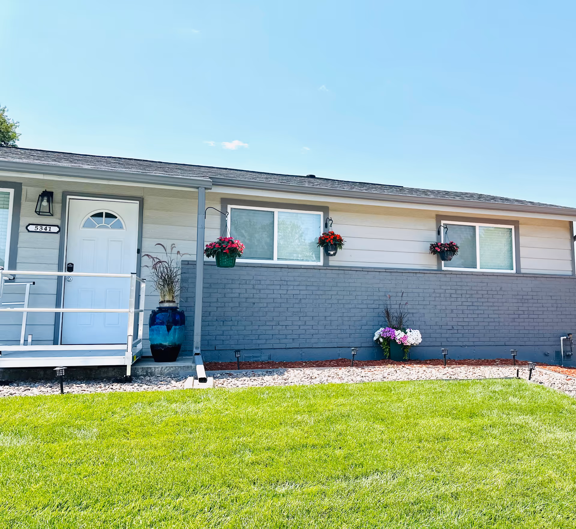 Single-story facility exterior with white and gray siding, windows, front door, and hanging planters.
