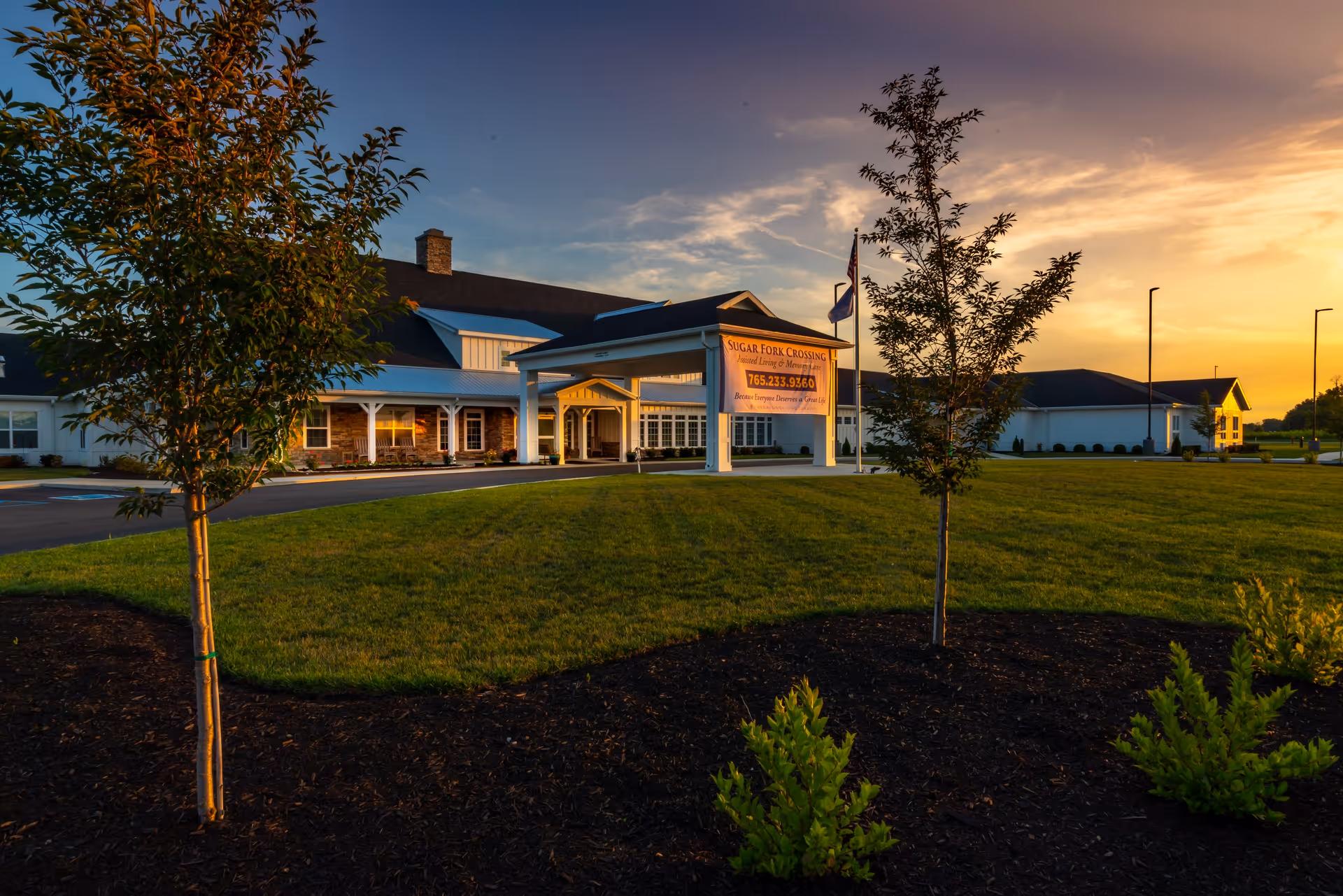 Exterior view of Sugar Fork Crossing senior living facility at sunset, showing a large building with a covered entrance, a green lawn, young trees, and a clear sky with warm lighting.