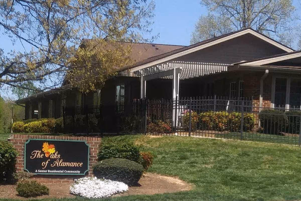 Front view of The Oaks of Alamance senior residential building with a lawn, entry porch, fence, and a sign.