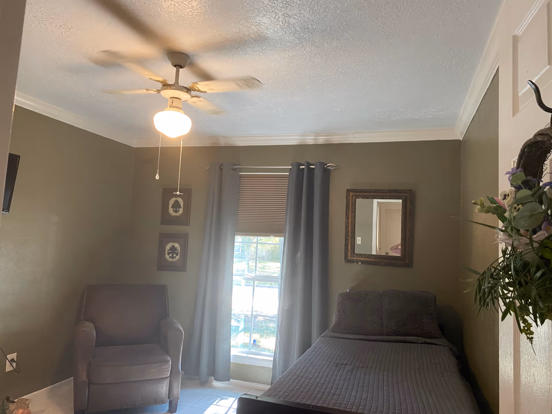 A small bedroom with a single bed, an armchair, window, curtains, and a ceiling fan.