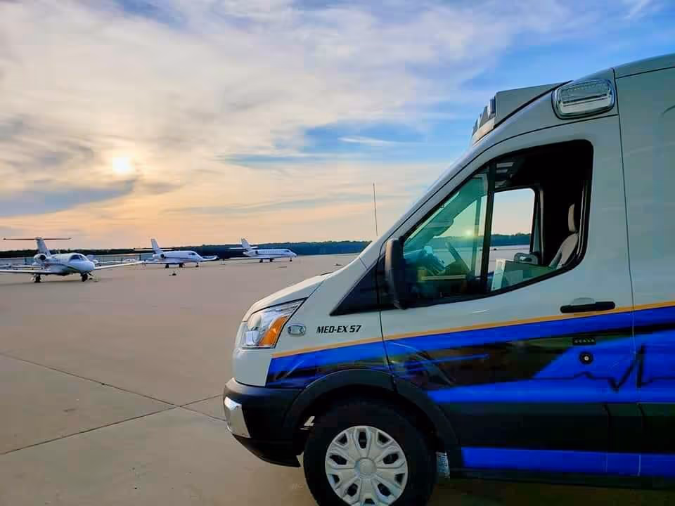 White medical transport van with blue stripe on tarmac beside small airplanes.