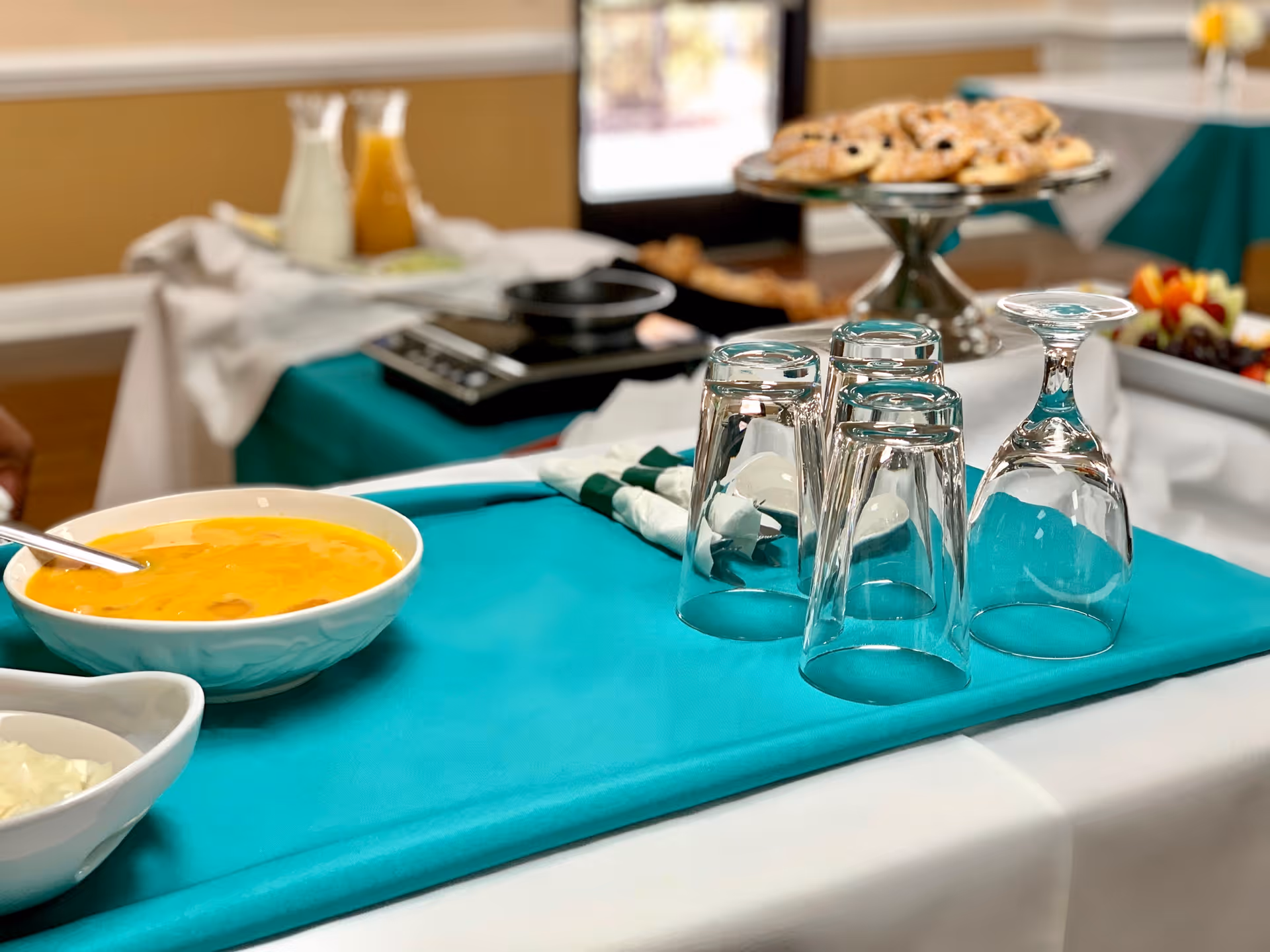A table set with upside-down drinking glasses on a teal tablecloth, a bowl of orange soup with a spoon, and a tray of pastries in the background inside a dining area.