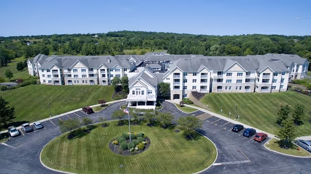 Aerial view of Clark Meadows senior living facility showing a large, multi-wing building surrounded by well-maintained green lawns, a circular driveway with a landscaped center island featuring a flagpole, and several parked cars. The background includes a tree-lined horizon under a clear blue sky.
