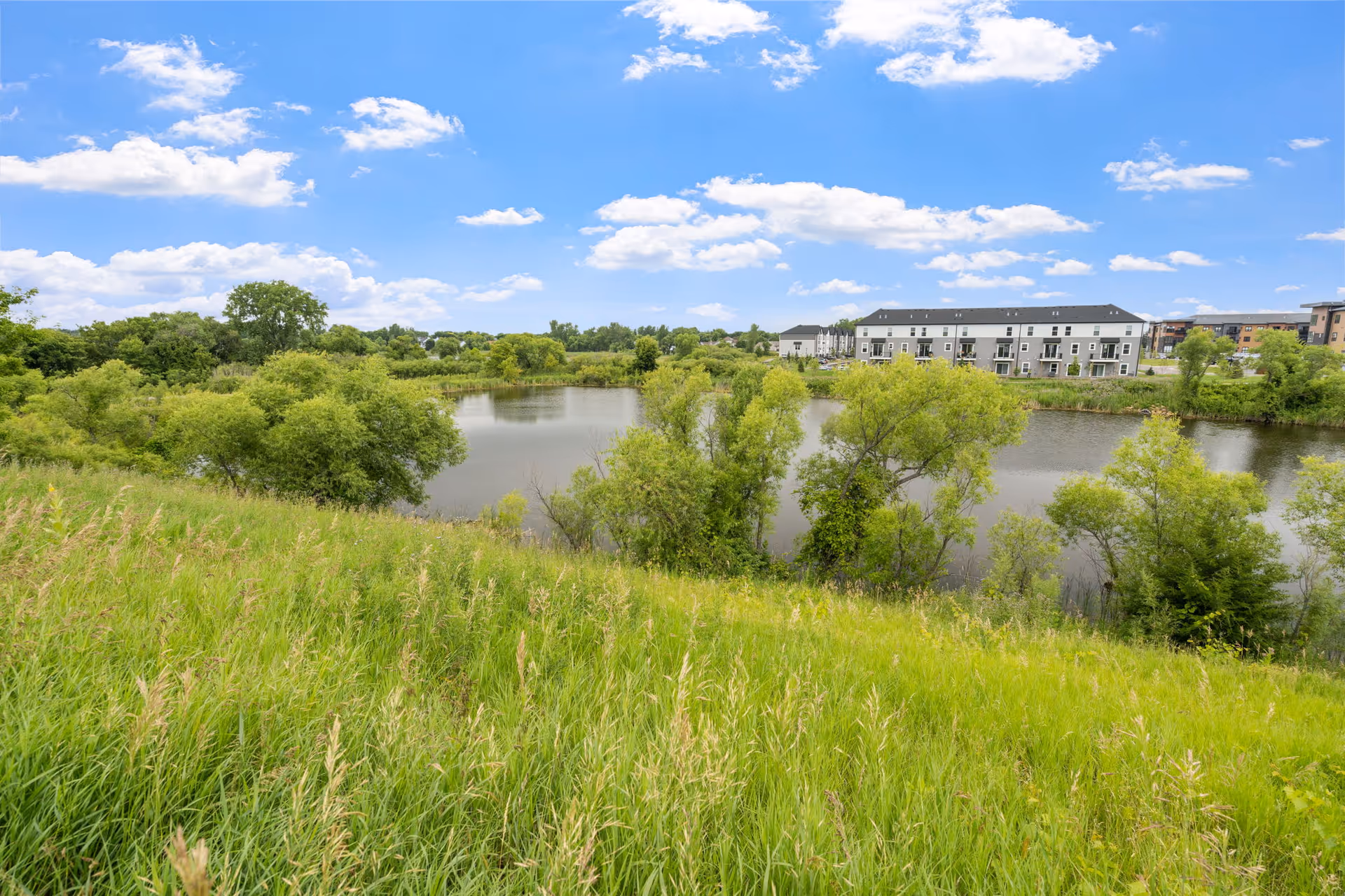 Outdoor landscape at NorBella Senior Living - Rogers with grassy banks, water, trees, and a building.