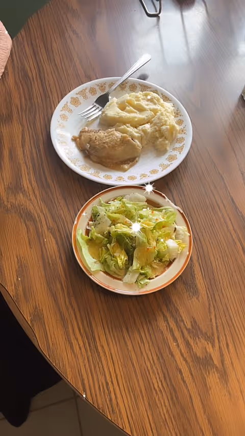 Two plates of food, including mashed potatoes, meat, and a green salad, on a wooden table.