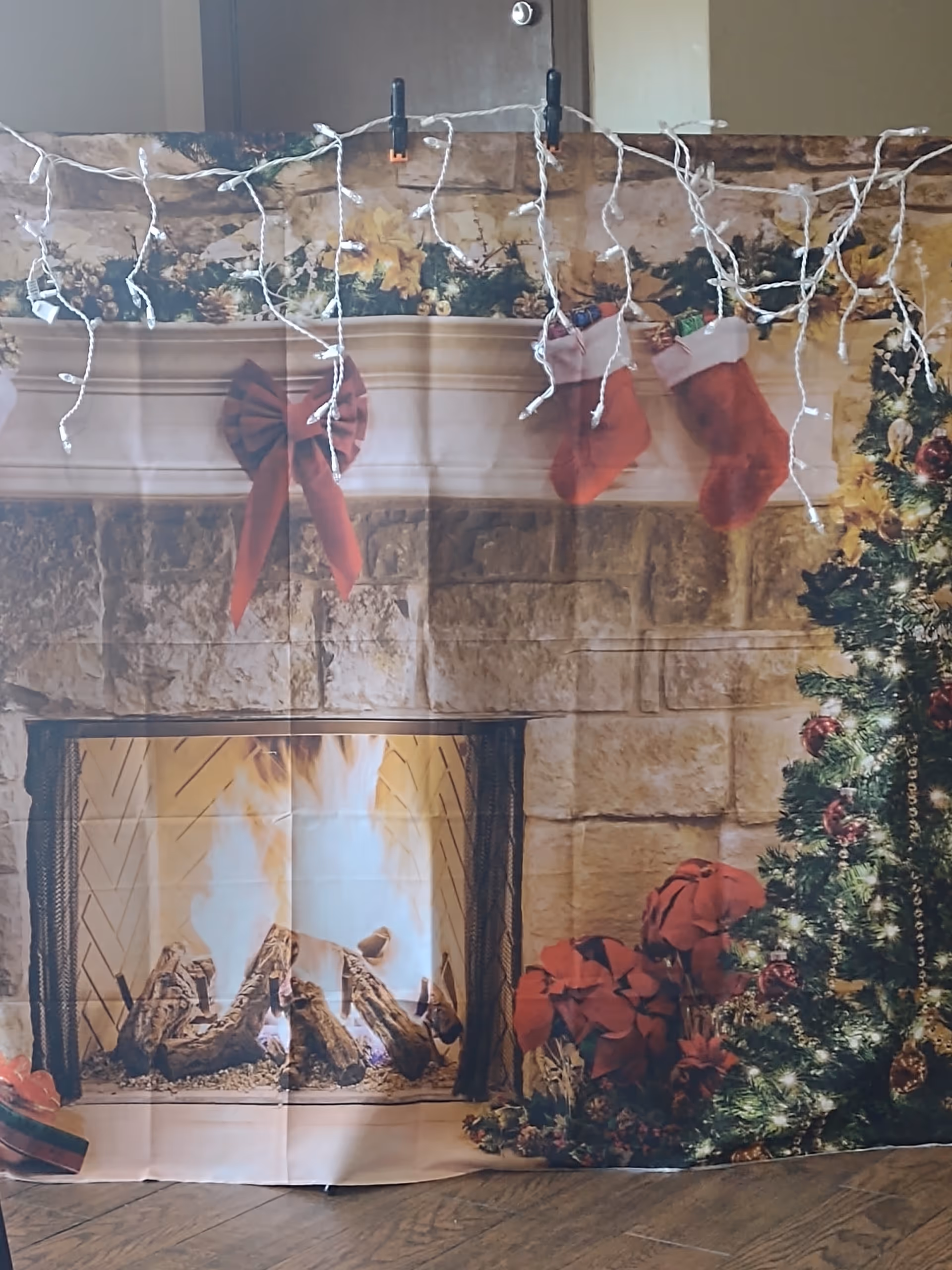 A decorative Christmas backdrop depicting a roaring fireplace, stockings on a mantel, a decorated tree, and string lights clipped across the top.