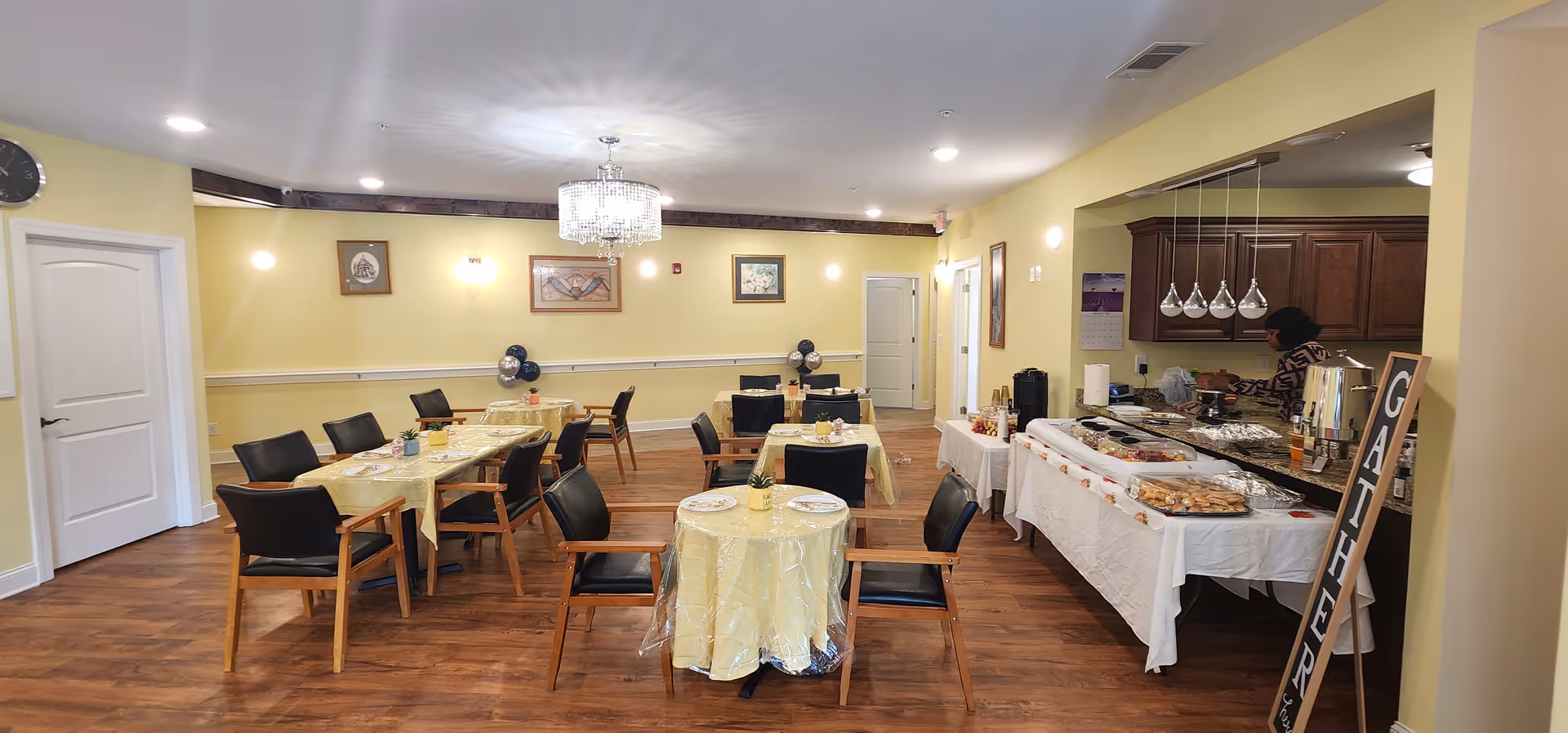 Bright dining hall with yellow walls, wooden floors, round tables, and a buffet area.