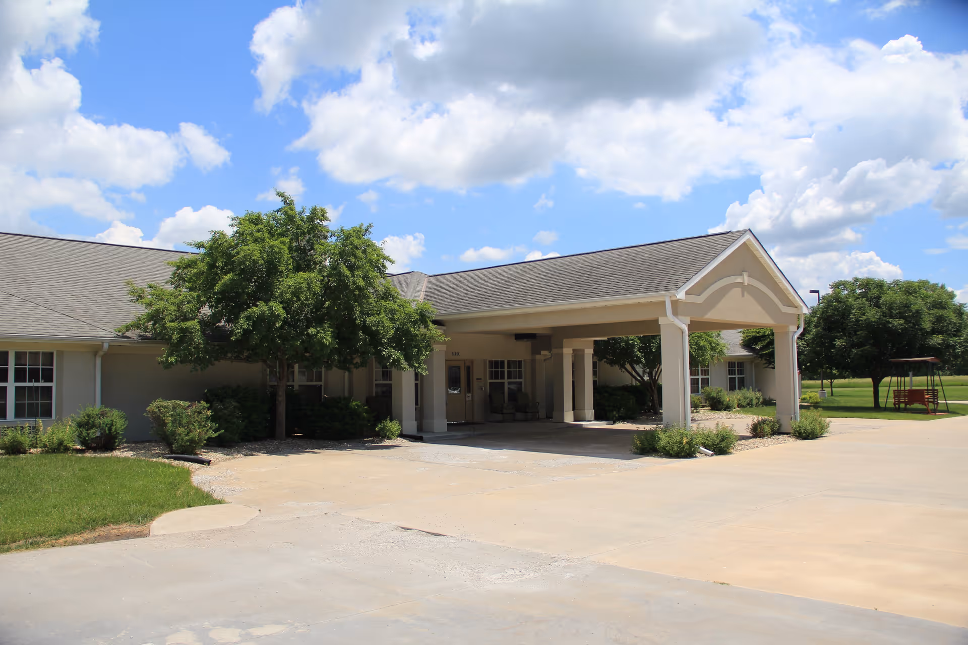 Exterior view of a single-story building with a covered entrance supported by columns, surrounded by green trees and bushes under a partly cloudy blue sky.