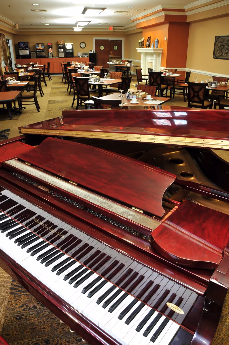 A polished grand piano in the foreground with a dining area in the background featuring multiple tables and chairs set for meals, a beverage station, and warm lighting in a senior living facility.