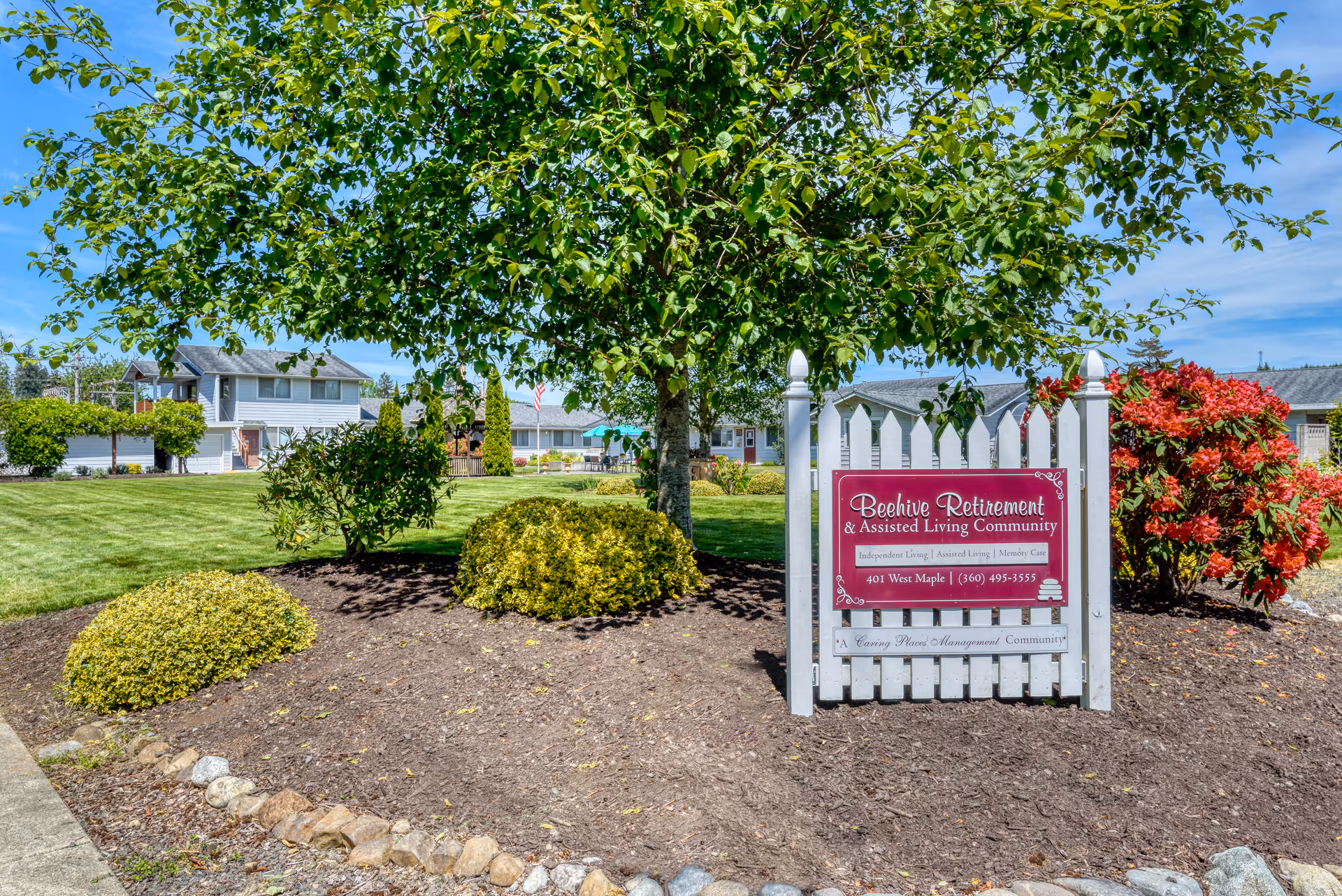 Landscaped front lawn with a white picket sign reading 'Beehive Retirement & Assisted Living Community' beneath a tree and buildings in the background.
