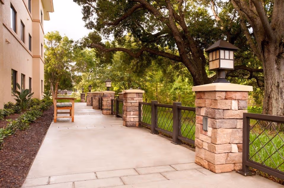 Outdoor walkway alongside a building with stone pillars topped with lantern-style lights, a wooden bench, and lush green trees and plants in the background.
