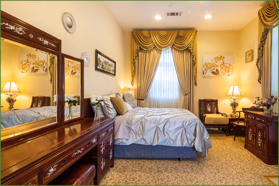 Traditional, well-lit bedroom with a made bed, ornate wooden dresser and mirror, bedside lamps, chairs, and draped curtains.