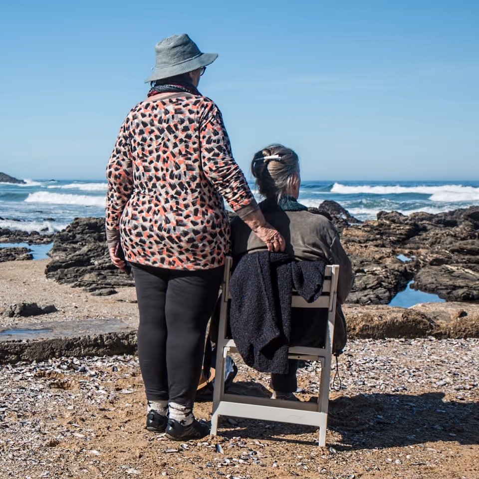 Two people, one standing, beside a seated elderly person, on a rocky beach by the ocean.