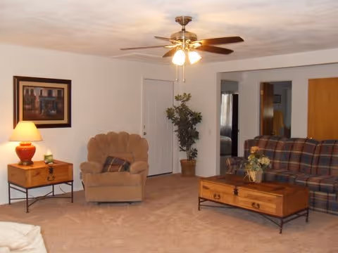 Cozy living room in a senior facility with sofa, chair, coffee table, lamp, and ceiling fan.