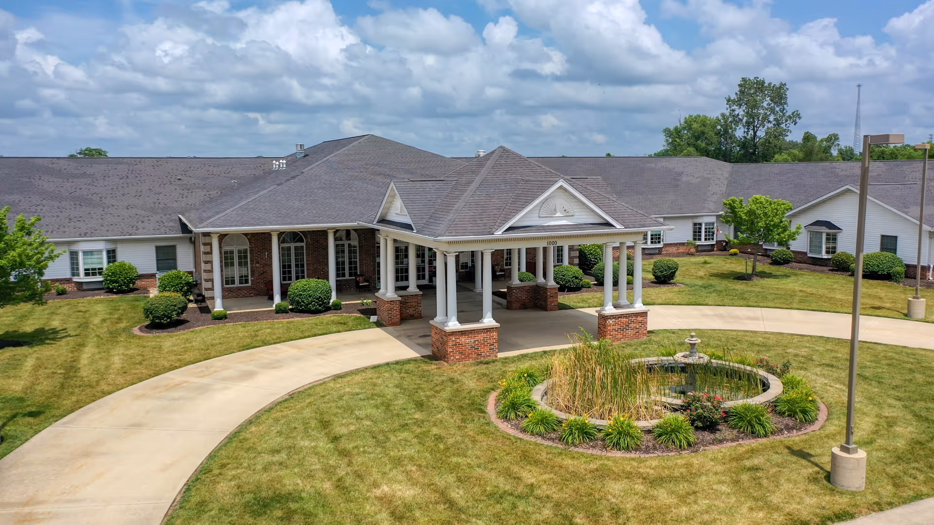 Front exterior view of Cedarhurst Senior Living of Godfrey showing a single-story building with a covered entrance supported by white columns and brick bases. The building is surrounded by well-maintained lawns, shrubs, and a circular driveway with a small landscaped fountain in the center. The sky is partly cloudy.