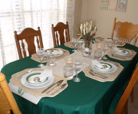 Dining table in a dining room set for four with a green tablecloth, place settings, glassware and a centerpiece.