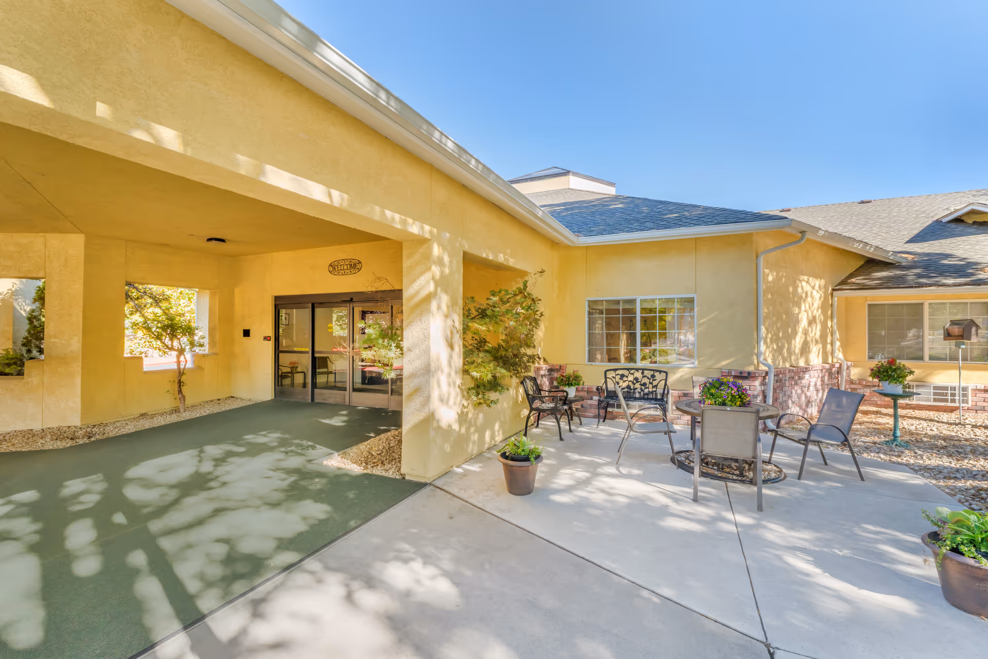 Entrance and outdoor patio seating area of a yellow-beige senior living facility with chairs, a table, and potted plants.