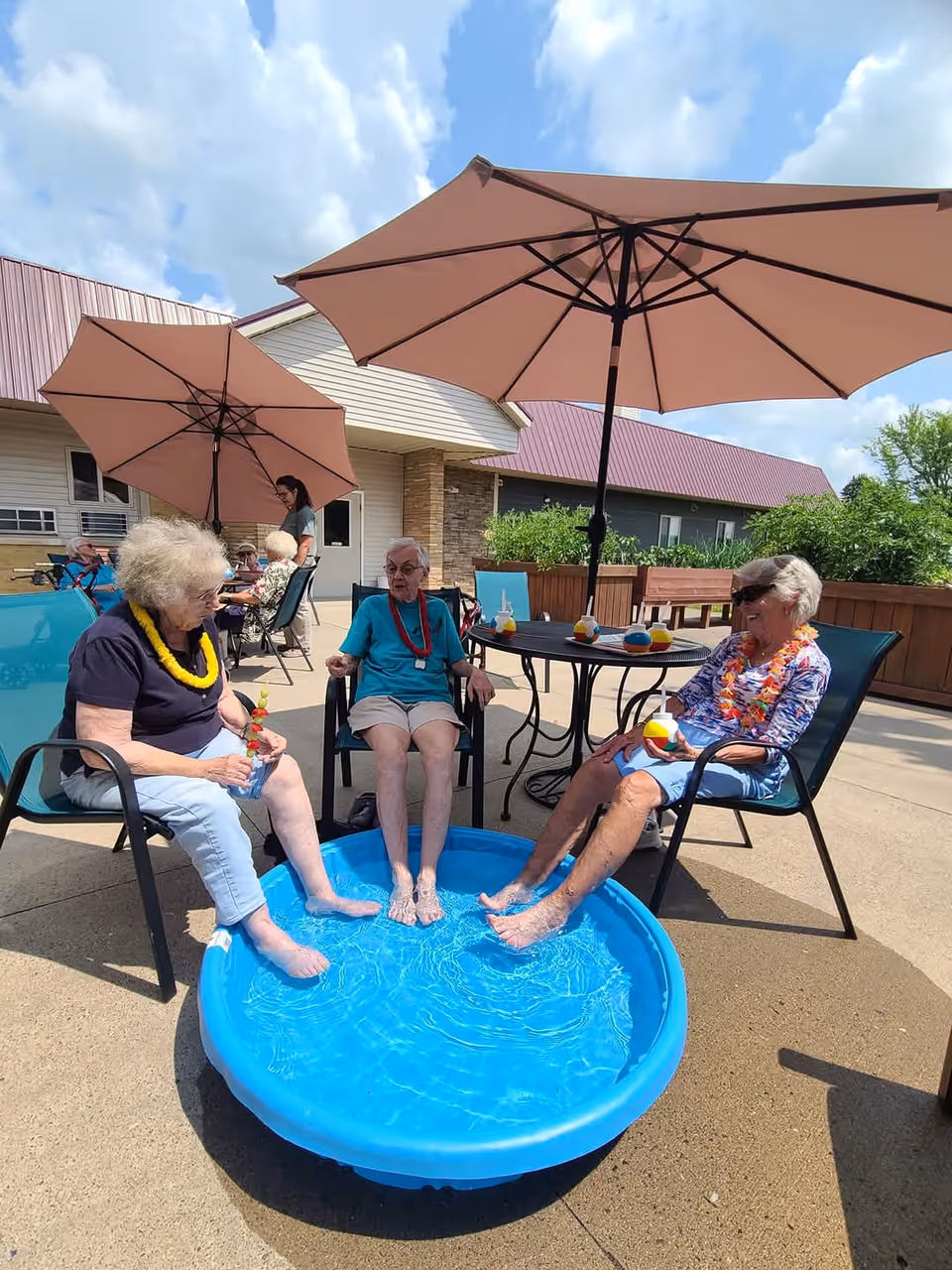 Three elderly women sit under patio umbrellas with their feet in a small blue wading pool on an outdoor patio at a senior living facility.