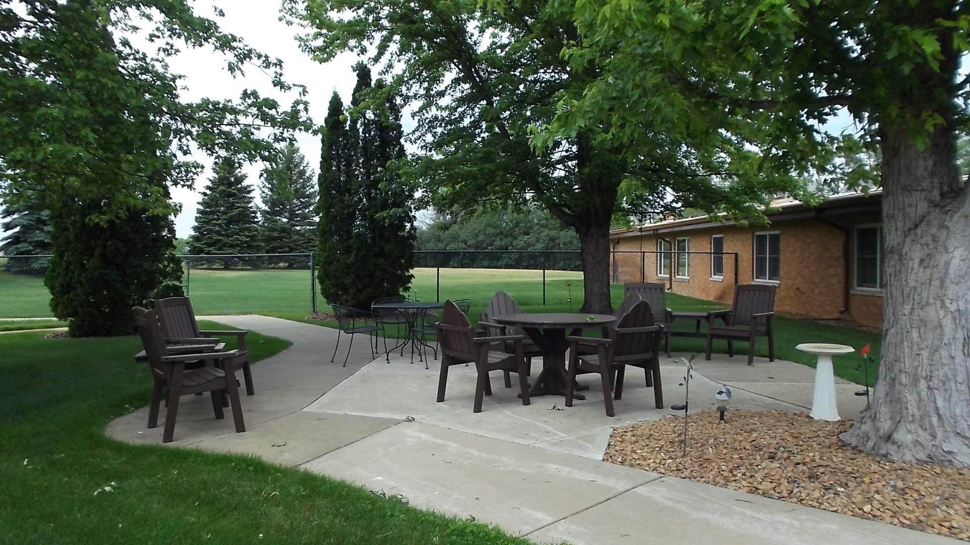 Outdoor patio area with several brown wooden chairs and tables arranged on a concrete surface surrounded by green grass, trees, and a brick building in the background.