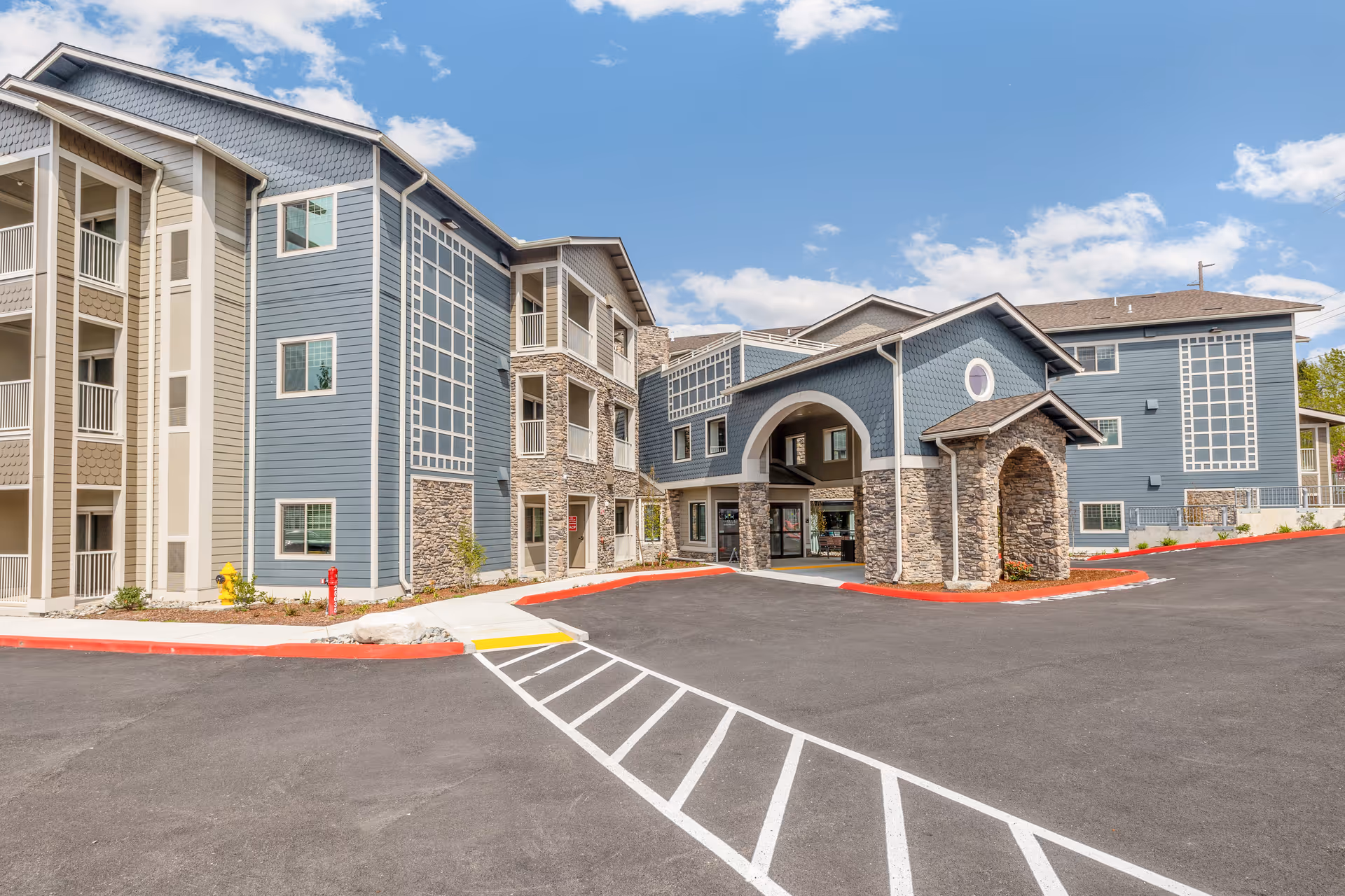 Exterior view of a modern senior living facility building with blue siding and stone accents under a partly cloudy sky. The building has multiple floors with balconies and a covered entrance area. The parking lot and driveway are visible in the foreground.
