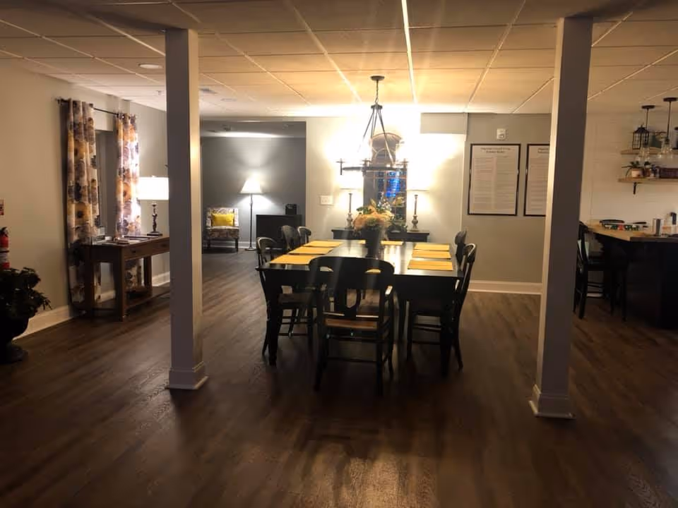 Interior dining room with a long wooden table and chairs under a chandelier, hardwood floors, and adjacent seating and console tables.