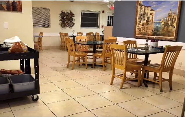 Interior view of a dining area with several wooden tables and chairs arranged on a tiled floor. A serving cart with trays and food items is visible on the left side. The walls are decorated with a large painting of a canal scene and a decorative wall piece. Windows and a door are seen in the background.