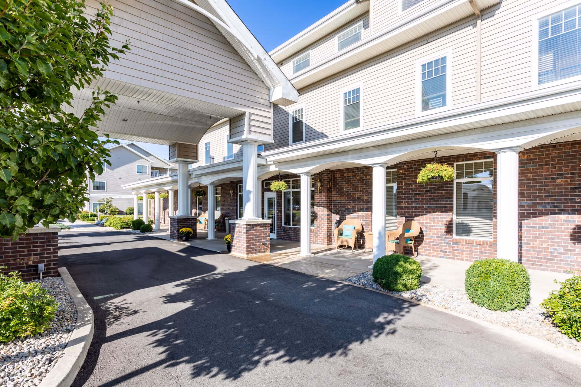 Exterior view of Broadway Court Estates showing a covered driveway entrance with white columns and brick walls. There are chairs with cushions on the porch area and hanging plants above. The building has multiple windows and is surrounded by neatly trimmed bushes and landscaping.