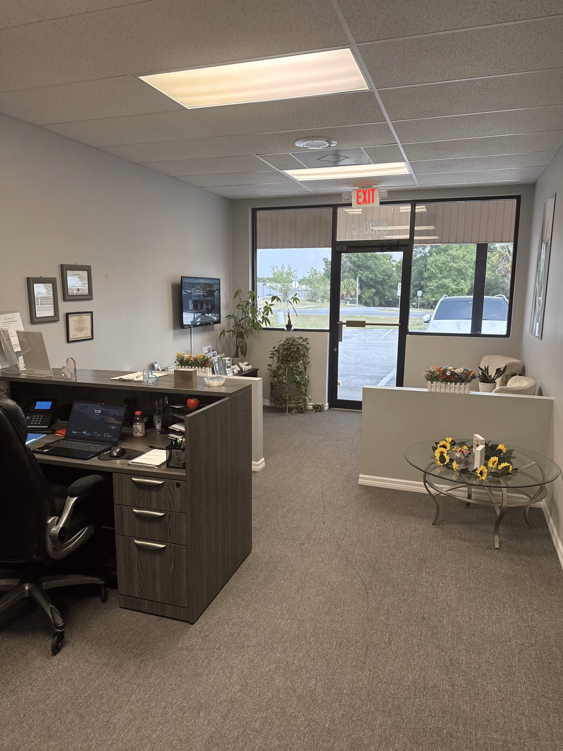 Office reception area with desk, chairs, plants, and a glass exit door.