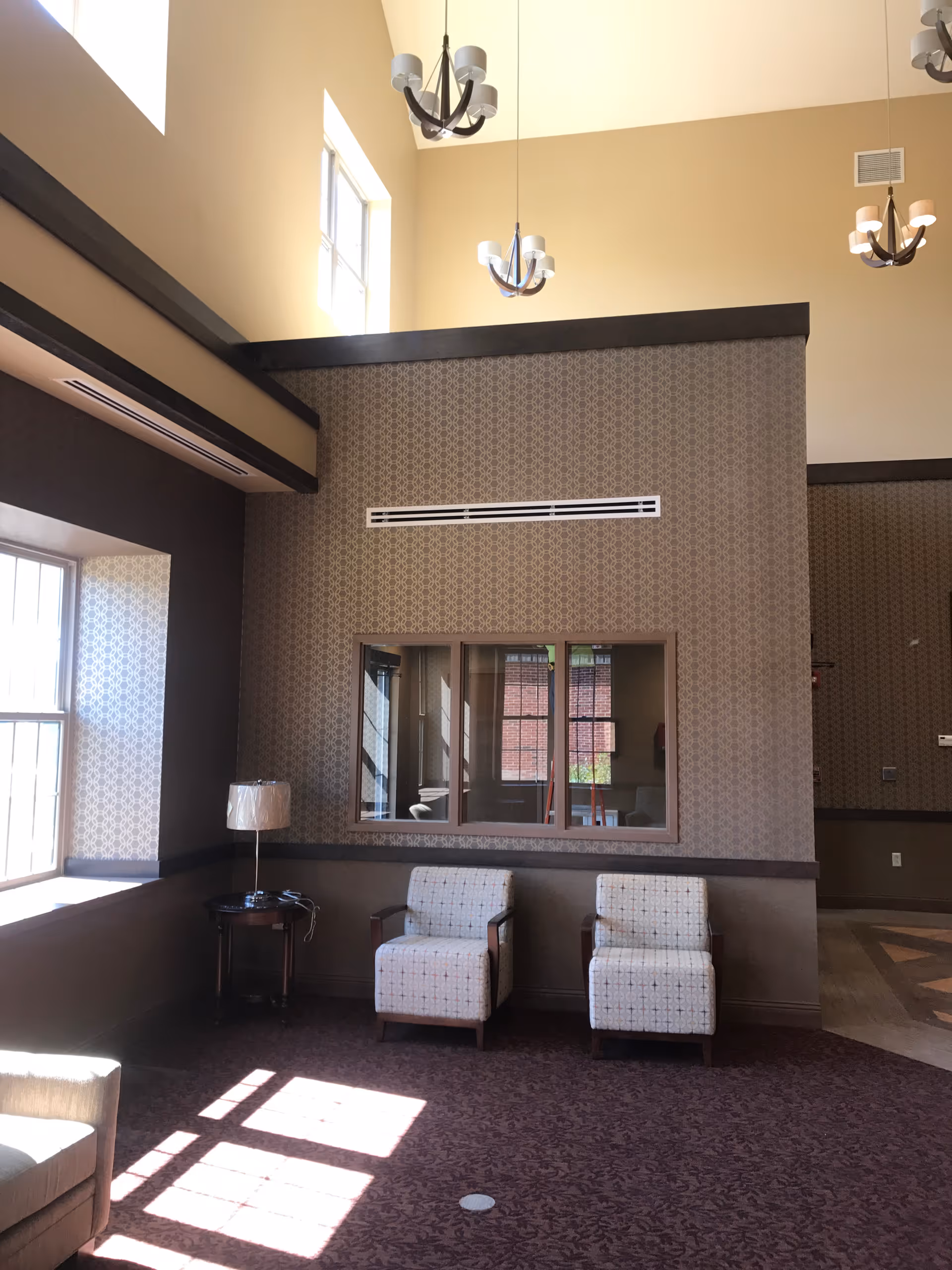 A tall-ceiling lobby with patterned walls, large windows, and two white patterned chairs.