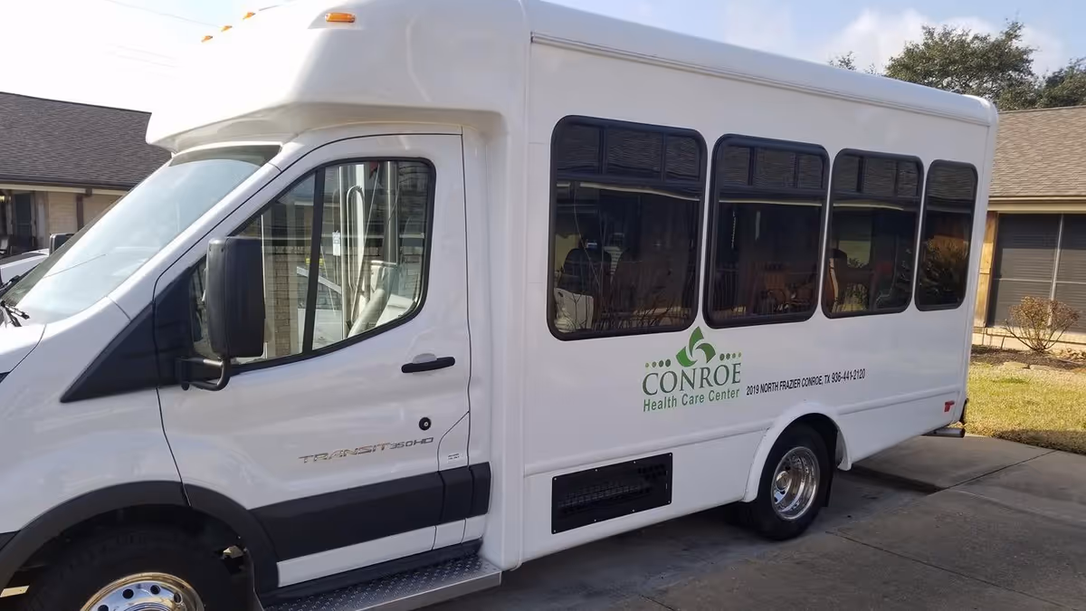 A white shuttle bus parked outside a building with the logo and text 'Conroe Health Care Center' on its side. The bus has large tinted windows and is positioned on a concrete driveway with grass and shrubs nearby.