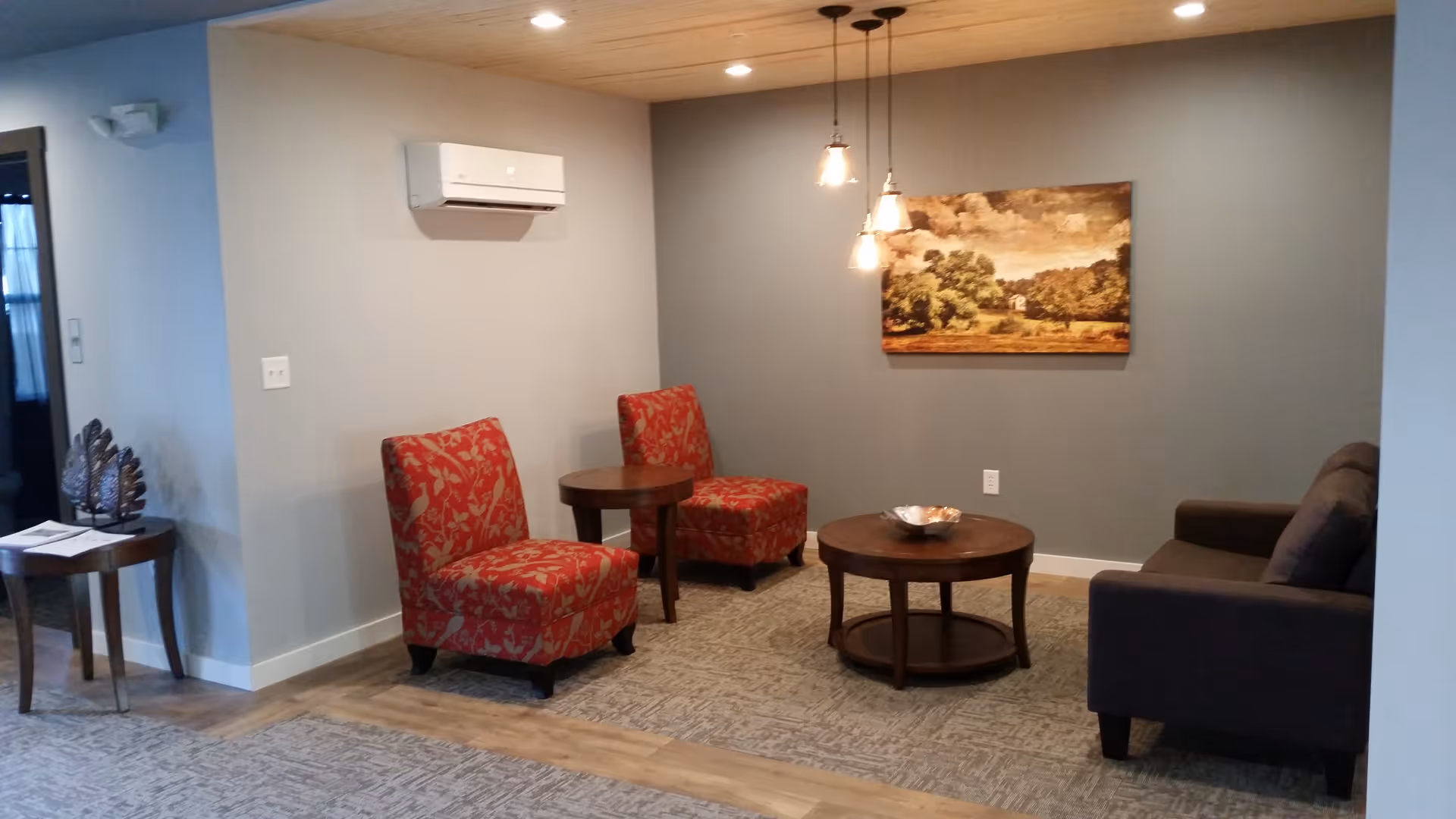 A cozy living room area with two red patterned armchairs, a dark brown sofa, a round wooden coffee table, and a side table. The walls are painted gray with a landscape painting hanging on one wall. The ceiling has wooden paneling with three pendant lights hanging down.