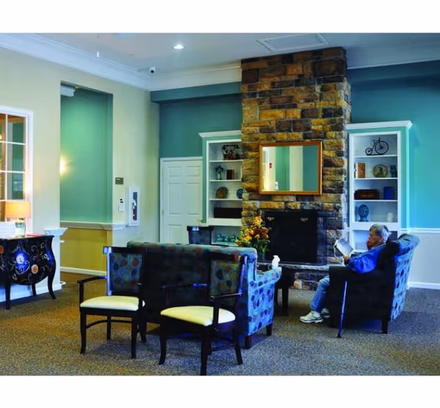 A lounge area with teal walls, a stone fireplace, built-in shelves, and a resident reading seated.