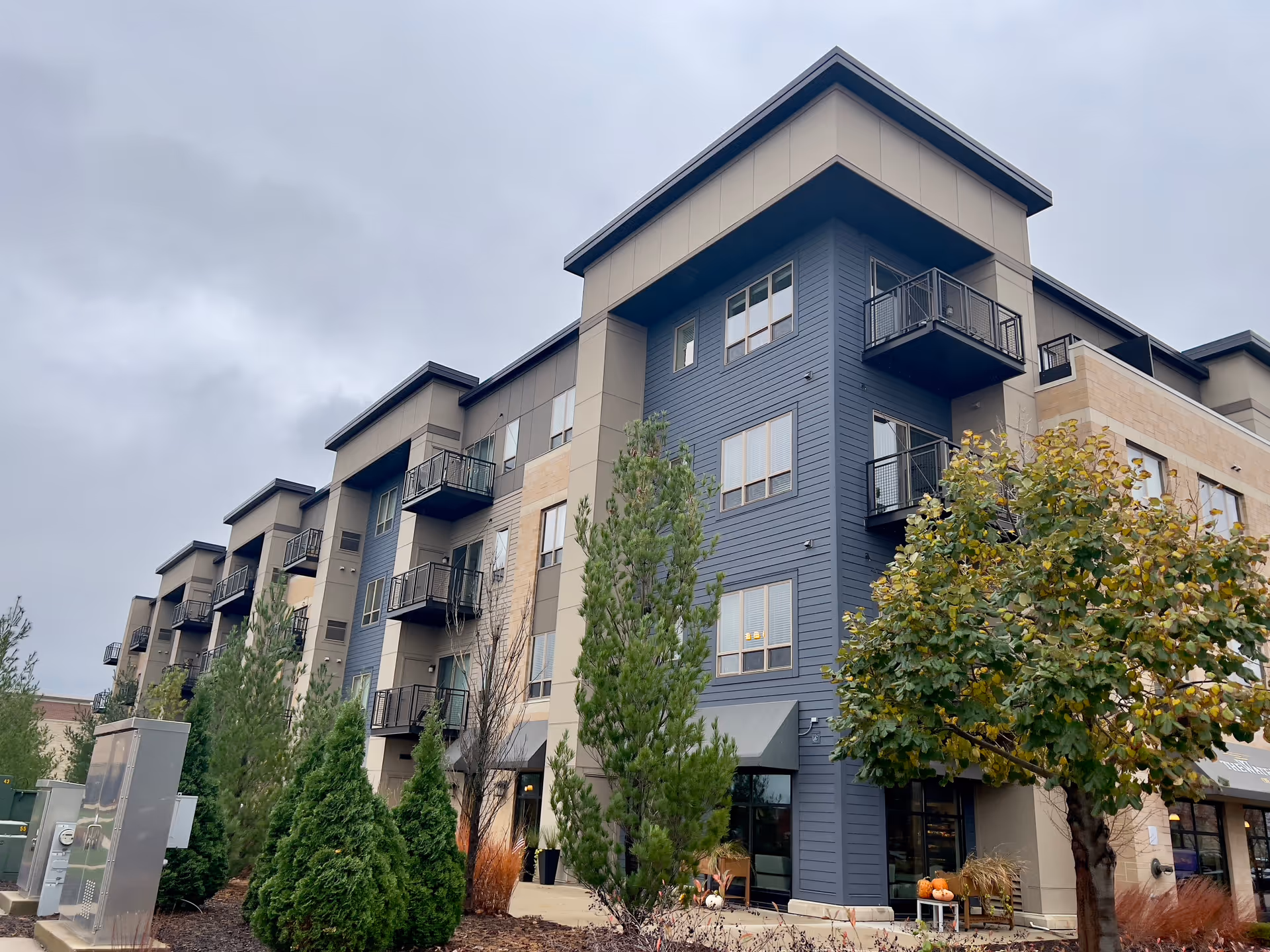 Modern four-story apartment-style building with balconies, trees, and a landscaped entrance under an overcast sky.