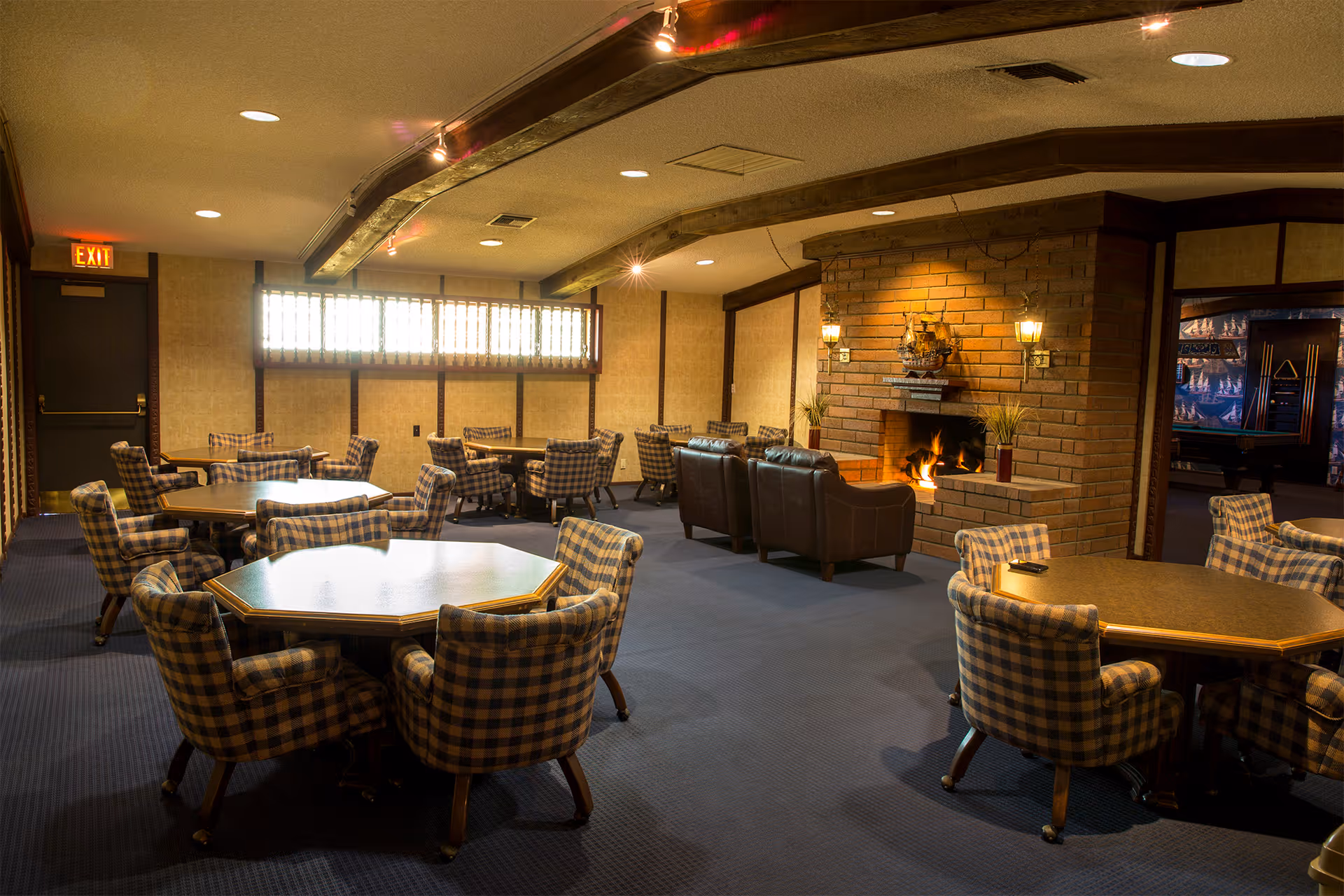 A cozy senior living community room with multiple hexagonal tables surrounded by plaid upholstered chairs. The room features a brick fireplace with a fire burning, flanked by two wall-mounted lamps and decorative items on the mantel. There are leather armchairs facing the fireplace and a pool table visible in an adjacent room. The room has carpeted flooring and warm lighting.